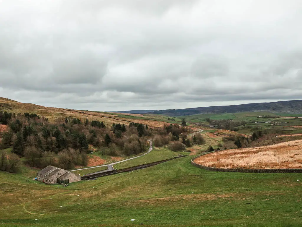 Looking down and across the underling fields, with a patch of woodland trees, and a small barn at the bottom of the hill.