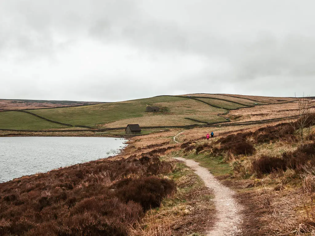 A narrow path winding down the side of the hill, with the water to the left, and a small barn ahead on the corner of the water. 