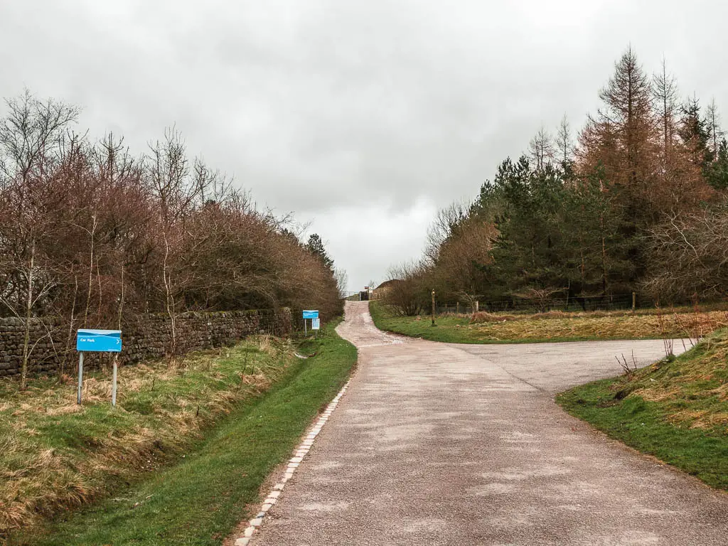 The road leading straight ahead and up, from the car path. There are two blue Yorkshire water signs on the left. There are woodland trees and a stone wall lining the road on the left, and a group of woodland trees ahead to the right. 