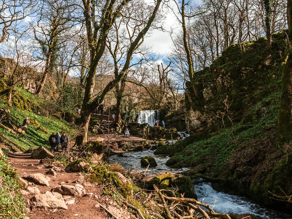 A rocky dirt path on the left and river on the right, and Janet's Foss waterfall visible ahead past some trees.