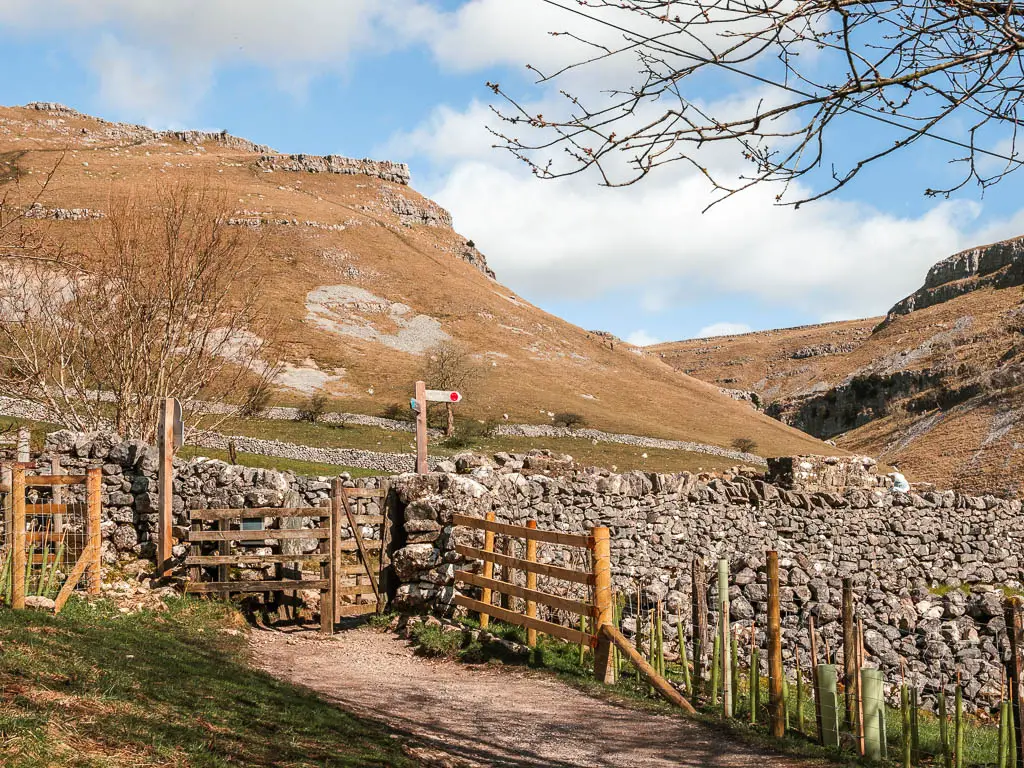 The path leading towards a wooden gate in the stone wall on the walk to Gordale Scar from Janet's Foss. There is a wooden trail signpost on the other side of the wall. There is a big hill rising up high ahead.