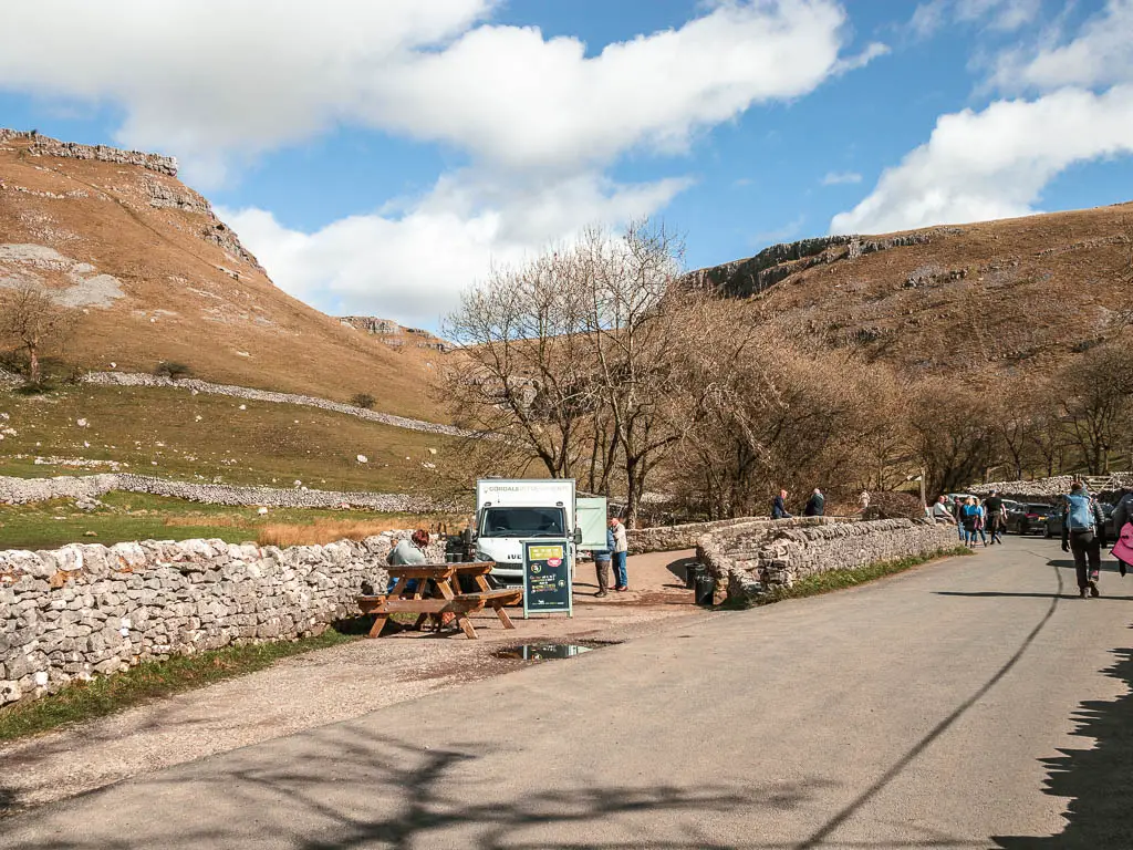 A road with a food truck to the left of it, and big hills behind it, on the walk towards Gordale Scar. There are lots of people walking about.