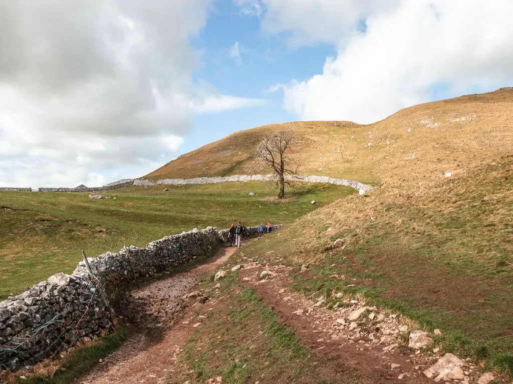 A rocky dirt trail through the hills, with a stone wall to the left. There are people walking ahead on the trail.