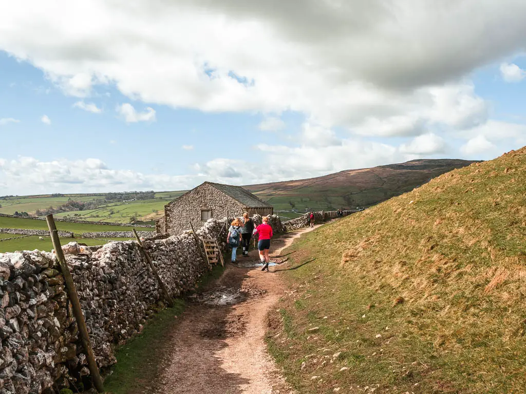 Lots of people waling on a dirt trail with a grass hill on the right, stone wall on the left, and a stone hut ahead on the left side of the wall.