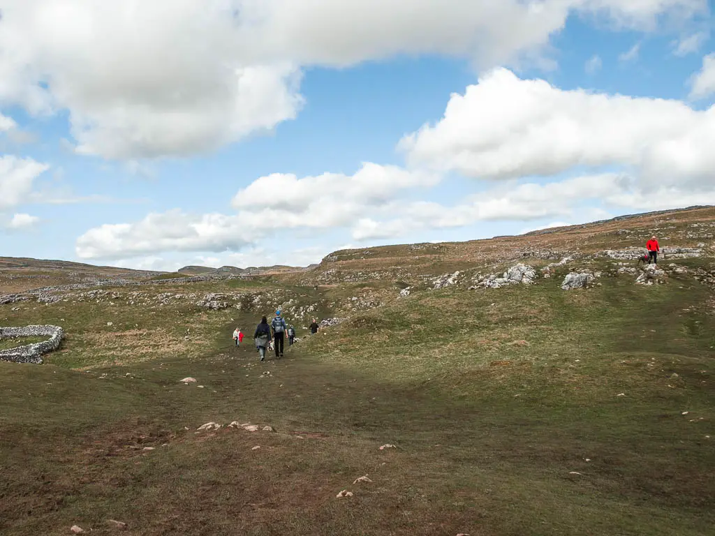 People walking cross the large grass hill field, with rocky bits ahead.