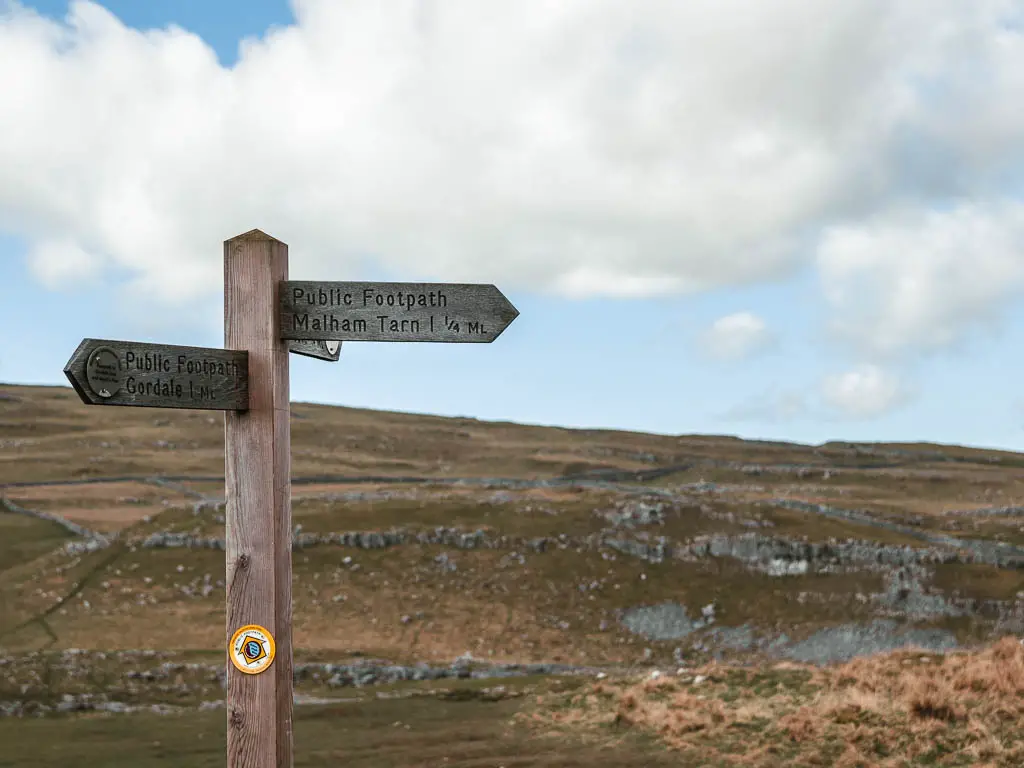 A wooden trail signpost pointing the way to walk to Malham Cove, Malham Tarn, and Gordale Scar.