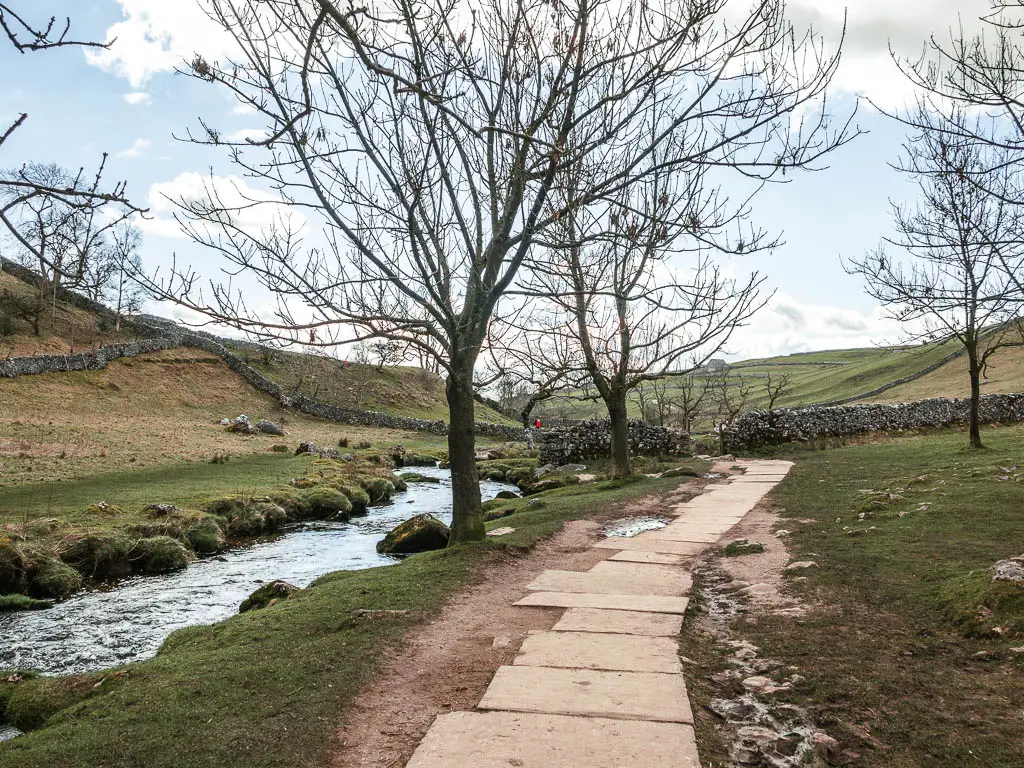 A stone paved path with a river on the left, on the walk away from Malham Cove. There are a few leafless trees next to the path.
