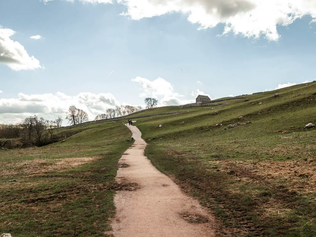 A long path leading straight through the grass field, heading slightly uphill. There is a small hut ahead on the top of the hill to the right.