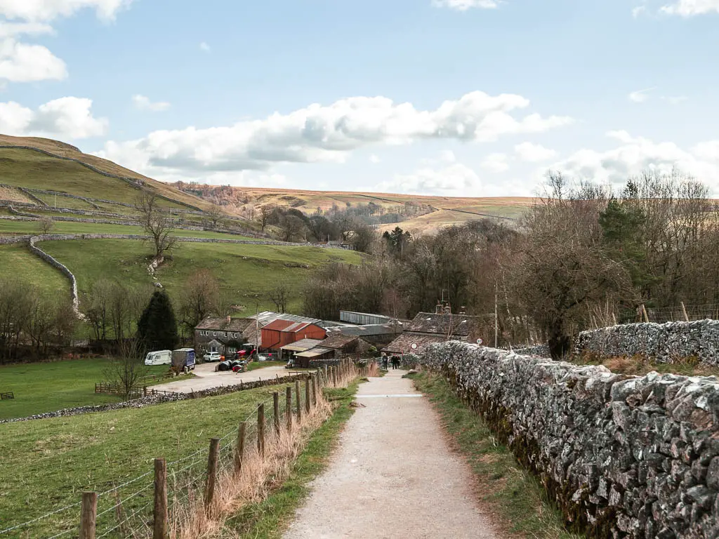 A wide path leading downhill to some farm buildings. There is a stone wall on the right of the trail, and wire fence on the left.