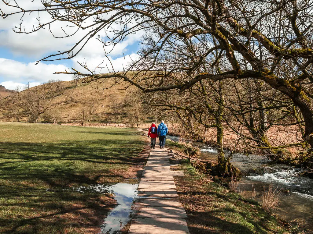Two people walking on a paved path, with the river on the right and field on the left, on the walk to Janet's Foss.