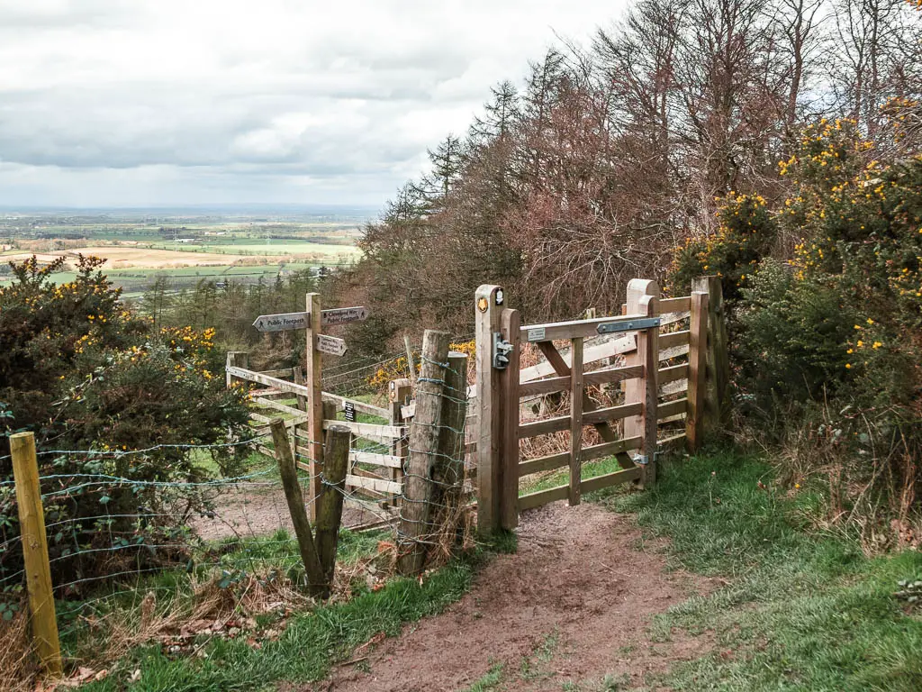 A wooden gate surround by bushes, and wooden trail signpost on the other side.