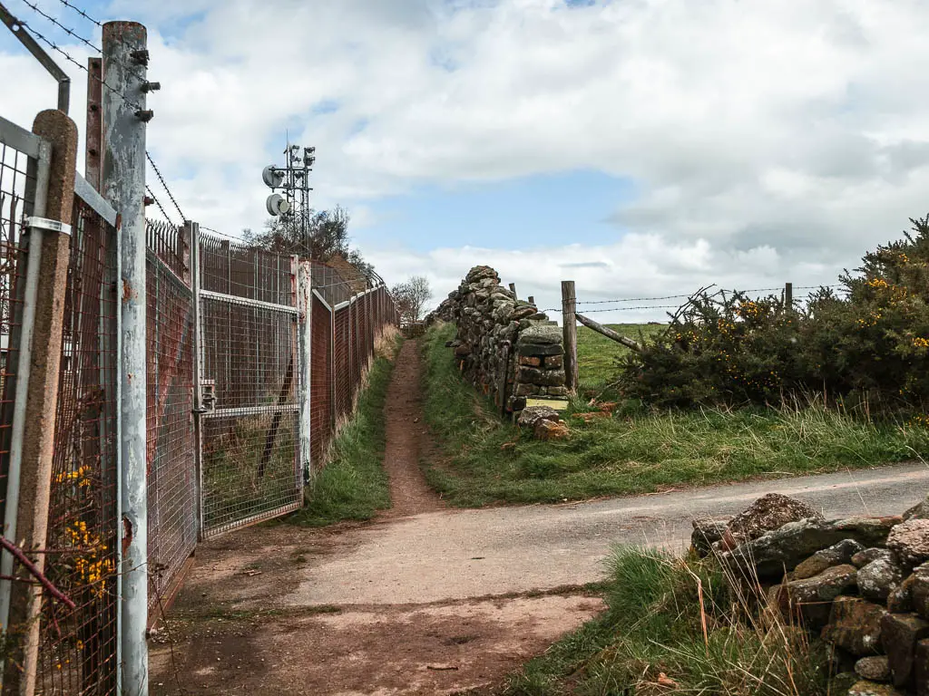 Metal gates and fence on the left and a dirt trial leading along side it straight ahead.