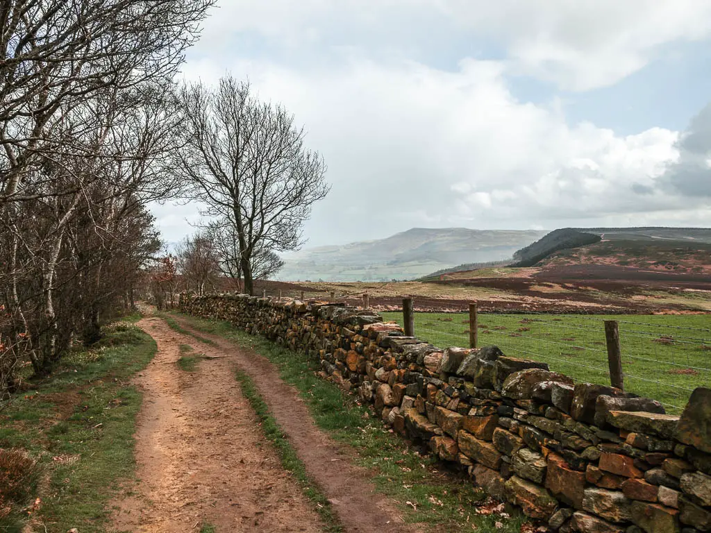 A dirt walking trail on the left, with a stone wall on the right side of it, along the Osmotherley circular route. There is a view to the hills in the distance.