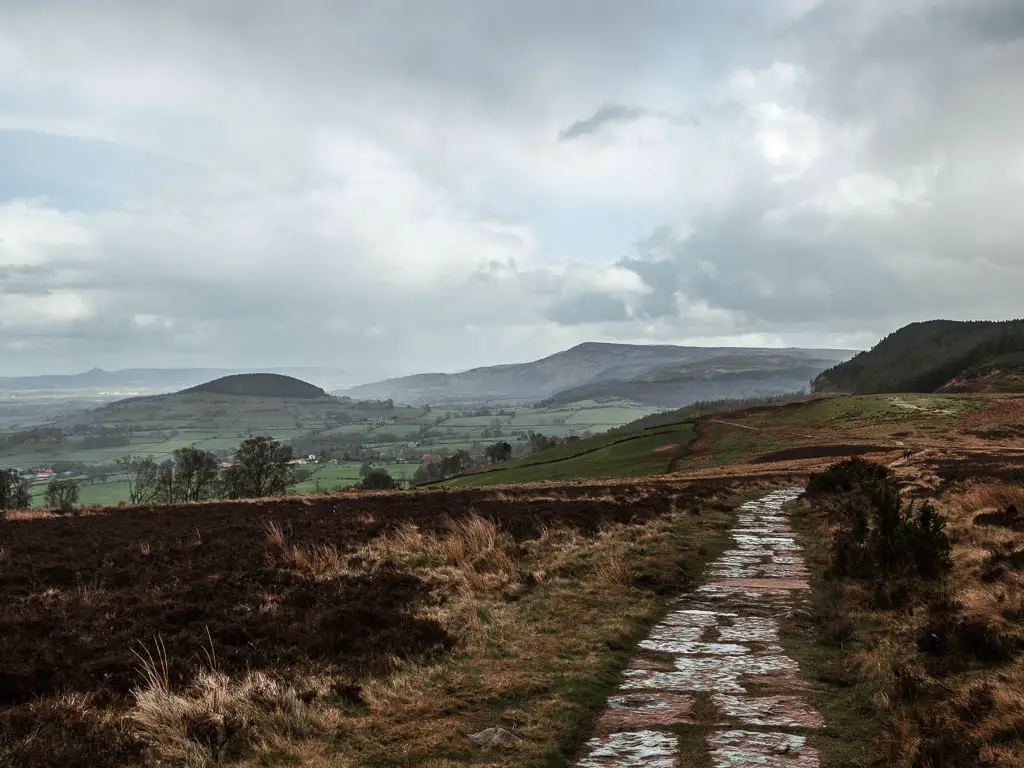 a paved trail on the right, through the Scarth Wood Moor, with the hills and fields stretching out into the distance on a rainy day.