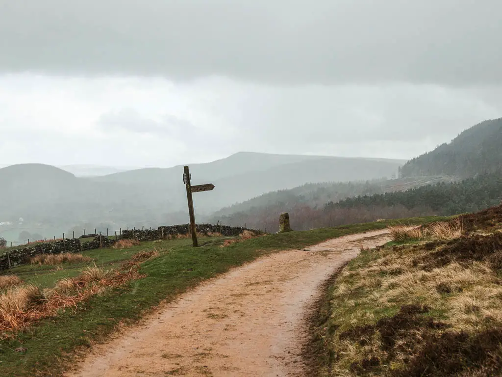 A path curving around to the right, with a wooden trail signpost pointing the way, and the silhouette of hills in the distance. 