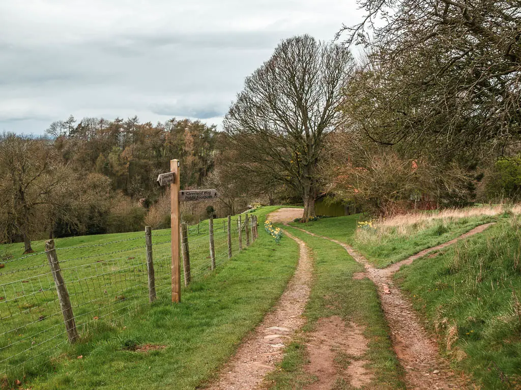 A wooden trail signpost on the side of a dirt track trail. There is a wire fence on the left of the sign with a green grass field on the other side.