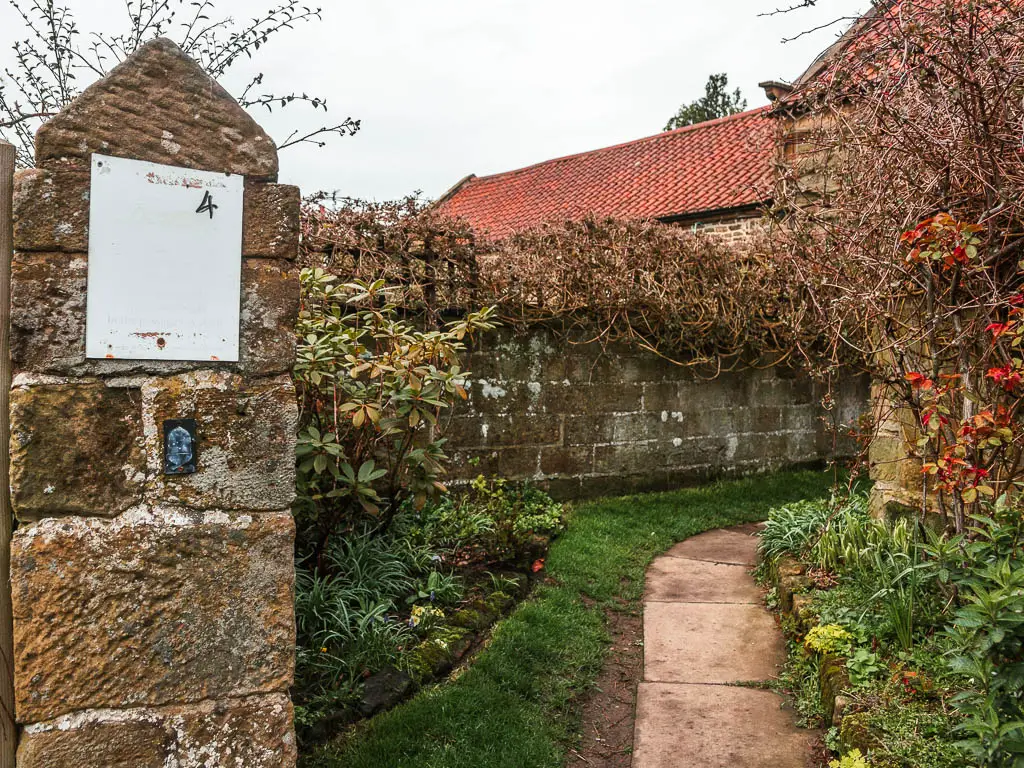 A very small black and white acorn sign on the stone pillars on the left, and a paved path leading through past the house on the right.