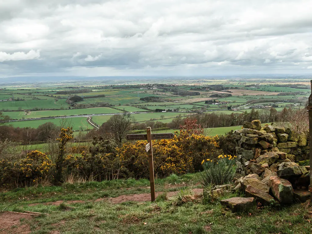 Looking down the hill to the view of the flat landscape of green fields. There is a wooden trail signpost just below.