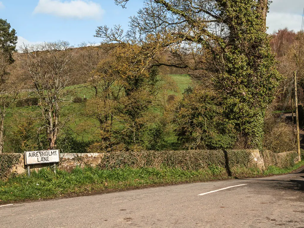 A road with stone wall covered in ivy along the side of it, and a road sign saying 'Aireyholme Lane'. there are lots of trees on the other side of the wall.