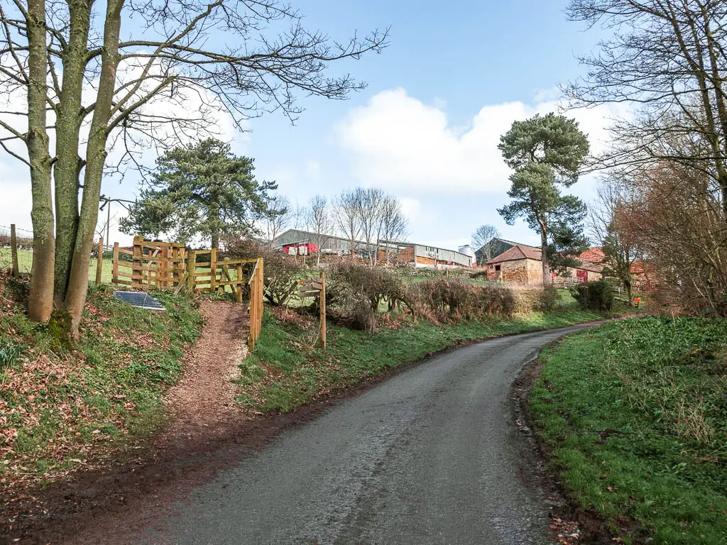The country road leading ahead to the right, with farm buildings ahead to the left, and a wooden trail signpost on the left side of the road pointing up to the wooden fence.