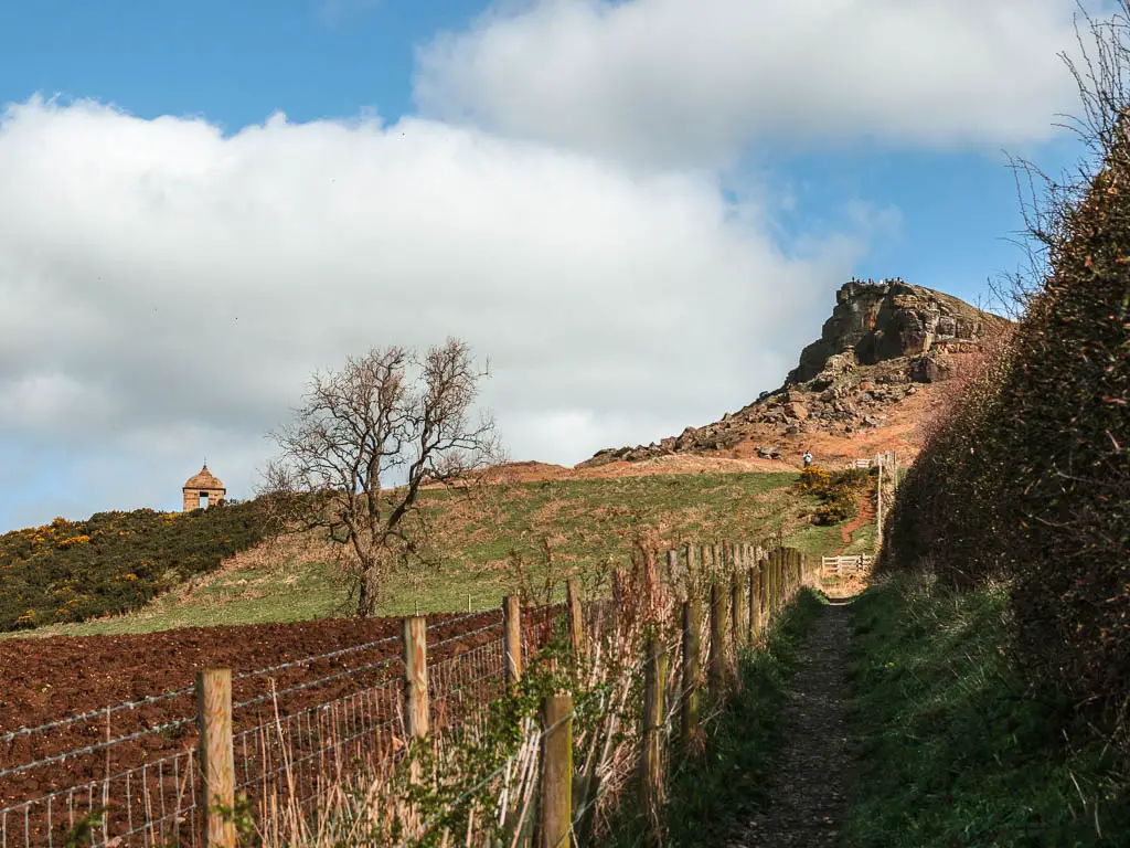 A narrow dirt trail on the right, leading towards the peak of Roseberry Topping, with a wire fence and crop field on the left.