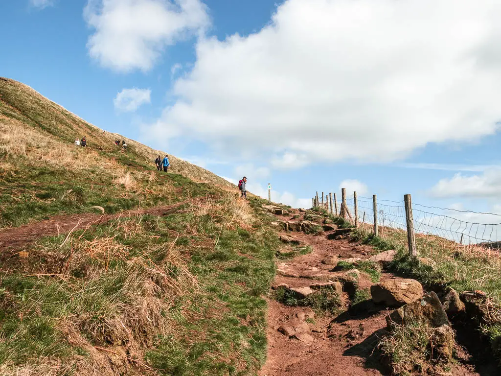A rugged and rocky dirt path on the right, with a grass hill on the left, on the walk up to the top of Roseberry Topping. There are a few people walking down the hill.