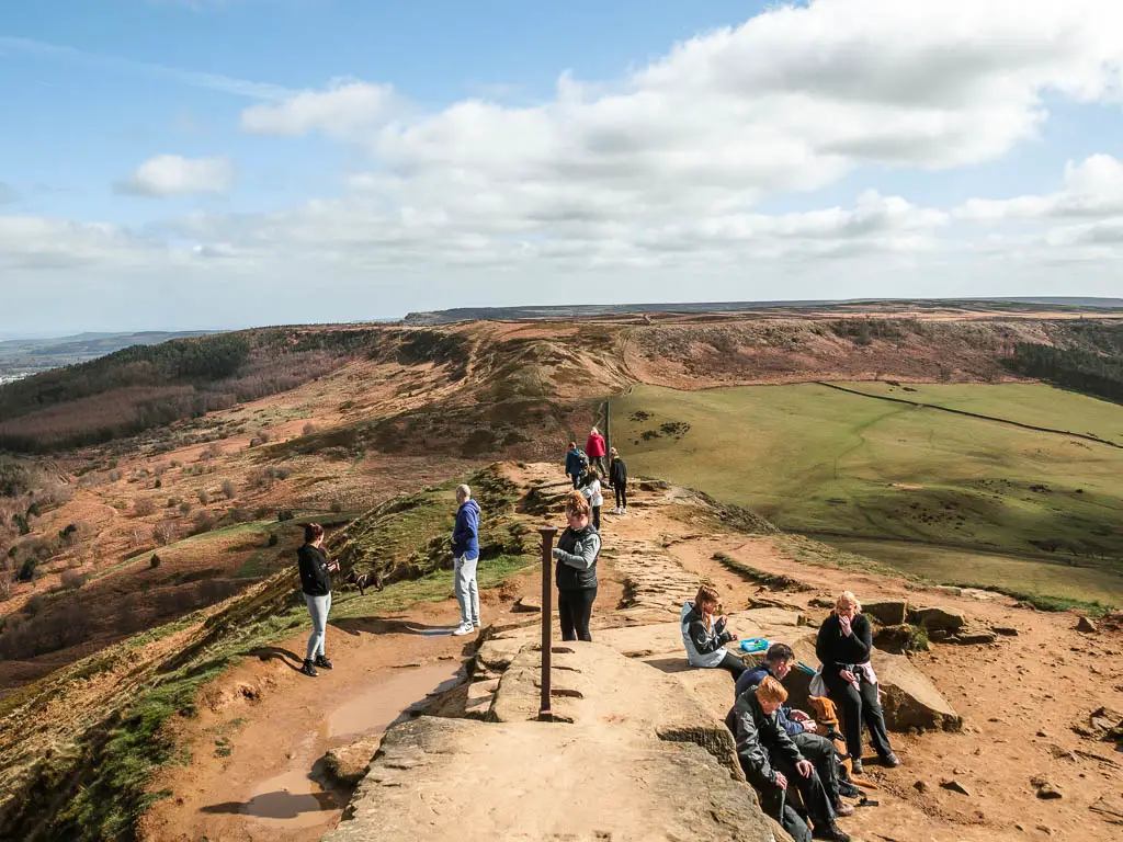 Lots of people sitting and walking around the summit of Roseberry Topping, with a view down into the valley and the ridge in the distance.
