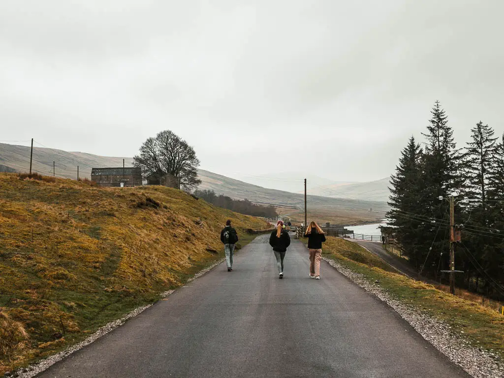 Three people walking along a road, with the Scar House Reservoir just about visible ahead to the right, and a hill to the left.