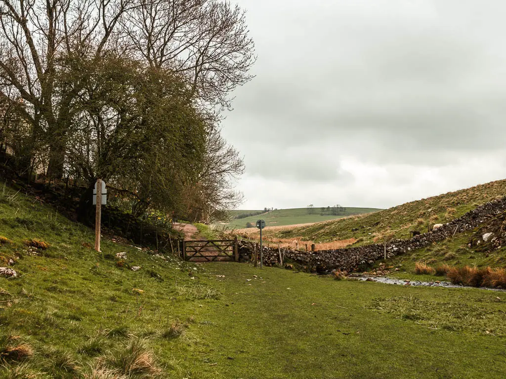 A wooden gate in the stone wall ahead, with a hill to the right, and trees to the left. 