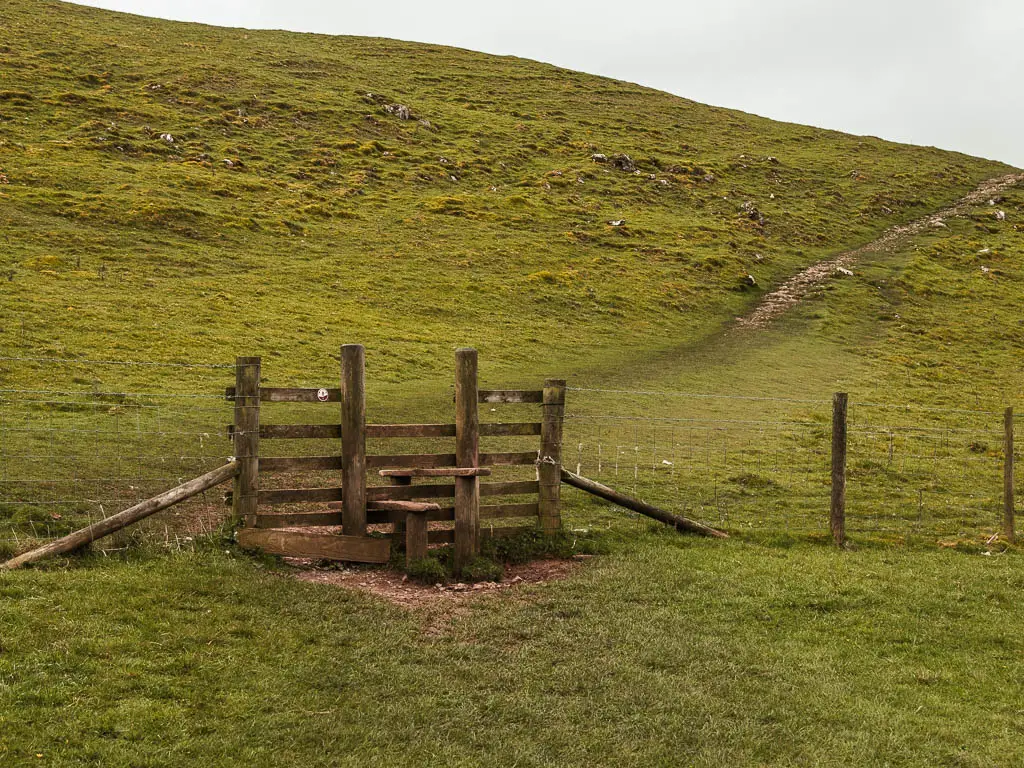 A wooden stile with a narrow trail leading up the hill on the other side.