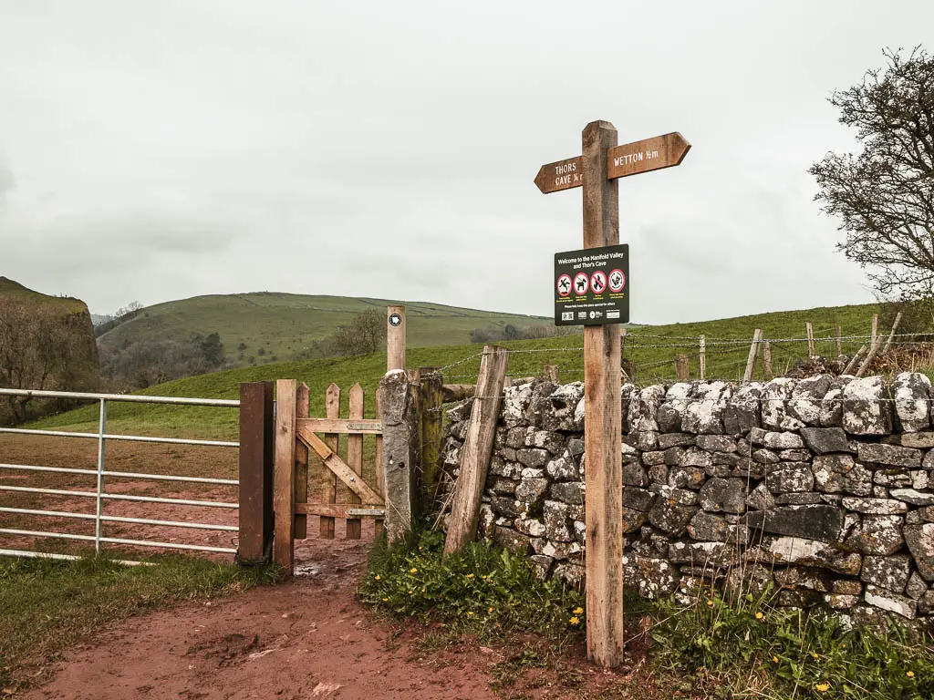 A wooden trail signpost next to a wooden gate and metal gait in the stone wall, pointing the way to walk to Thor's Cave.