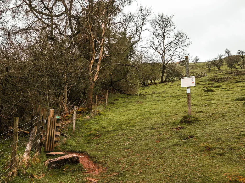 A wire fence and wooden gate on the left, and a trail signpost on the right, on the grass hillside. 