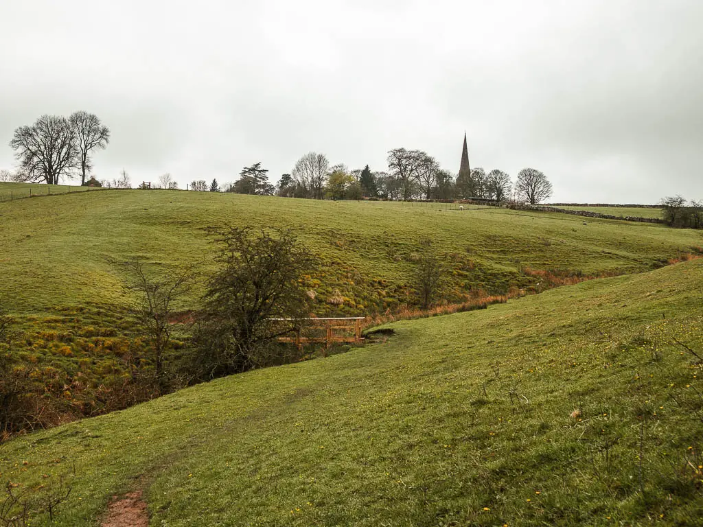 Looking down the grass hill to the dip, and a hill leading up the other side. There is a wooden bridge in the dip, and the church spire is visible ahead over the hill.