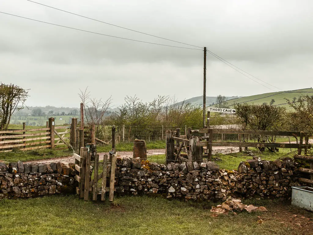 A small wooden gate in the stone wall, with a sign pointing right to walk tor Thor's Cave. There is a road on the other side of the wall, and a wooden gate and fence on the other side of the road.