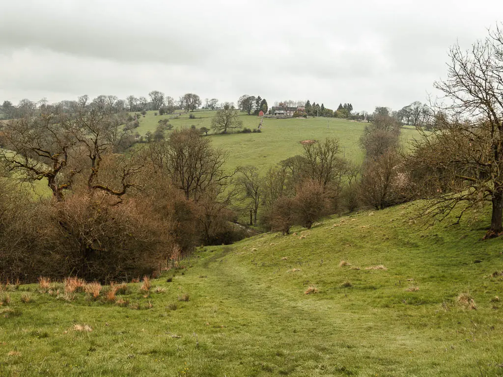 Looking down the grass hill, to the valley with trees below, and a hill rising up on the other side with a house on top.