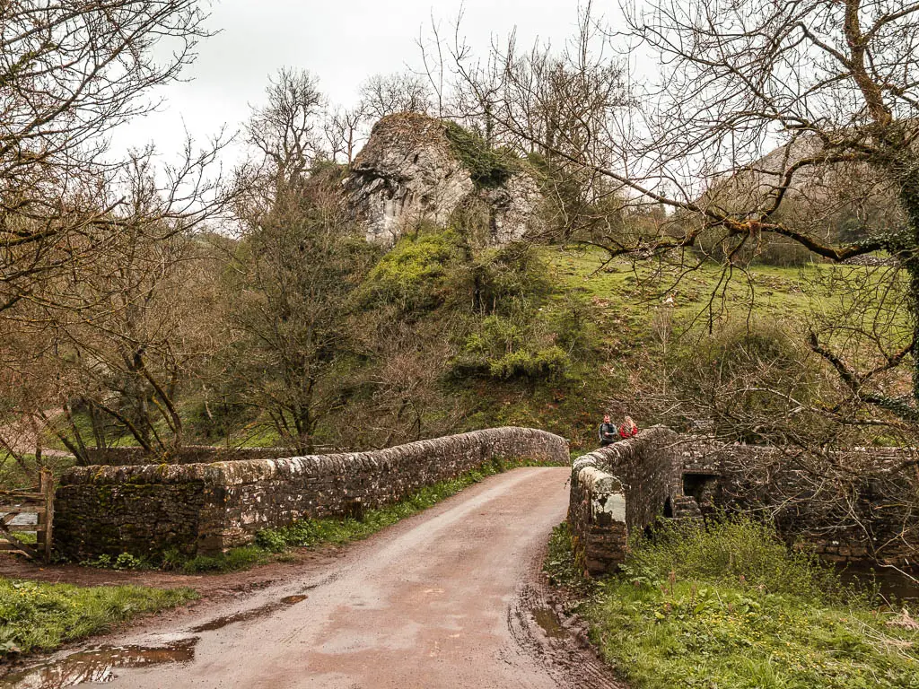 The road leading over a stone walled bridge with a cliff and hill on the other side. There are two people walking over the bridge.