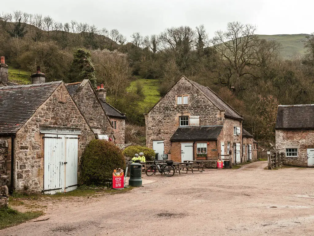 The stone buildings of Wetton Mills. There are wooden picnic benches outside, and a couple of cyclists sitting at them.