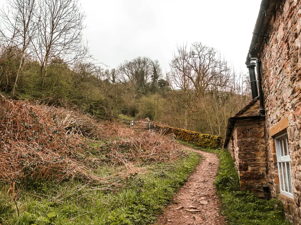 A rocky dirt trail leading uphill along the back of a stone walled cottage. 