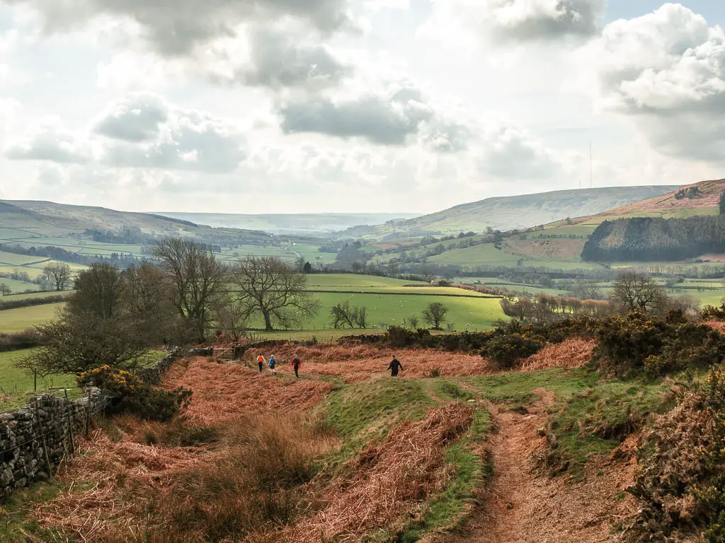 Looking down along the undulating hillside, to the valley and hills in the distance, on the walk towards the Wainstones. There are four people walking down the hill.