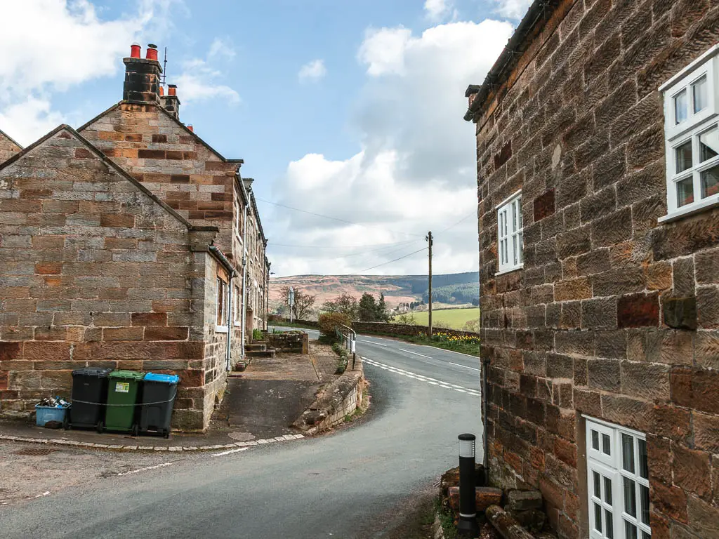 A stone walled house on the right, and some more on the left and a road through them leading to the main road. There are three rubbish bins outside the house on the left.