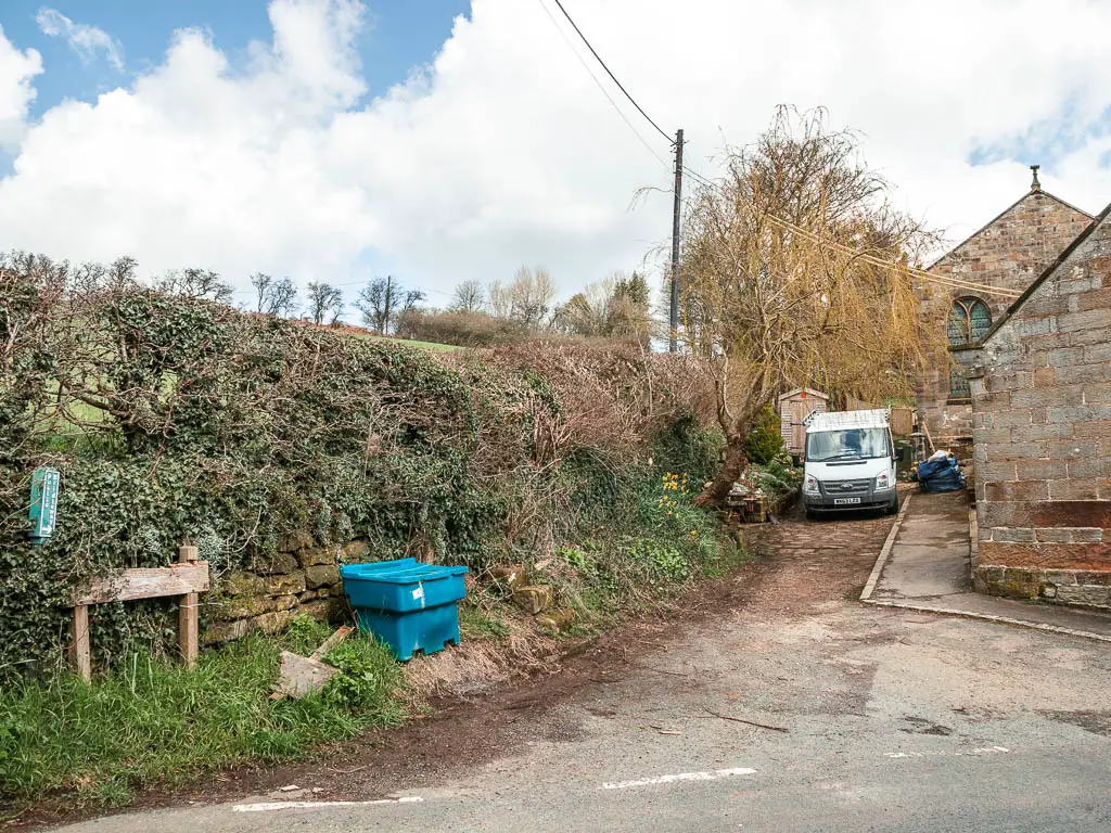 A small drive leading behind some houses, with a hedge on the left side of it and the houses to the right. There is a white van parked on the drive. 