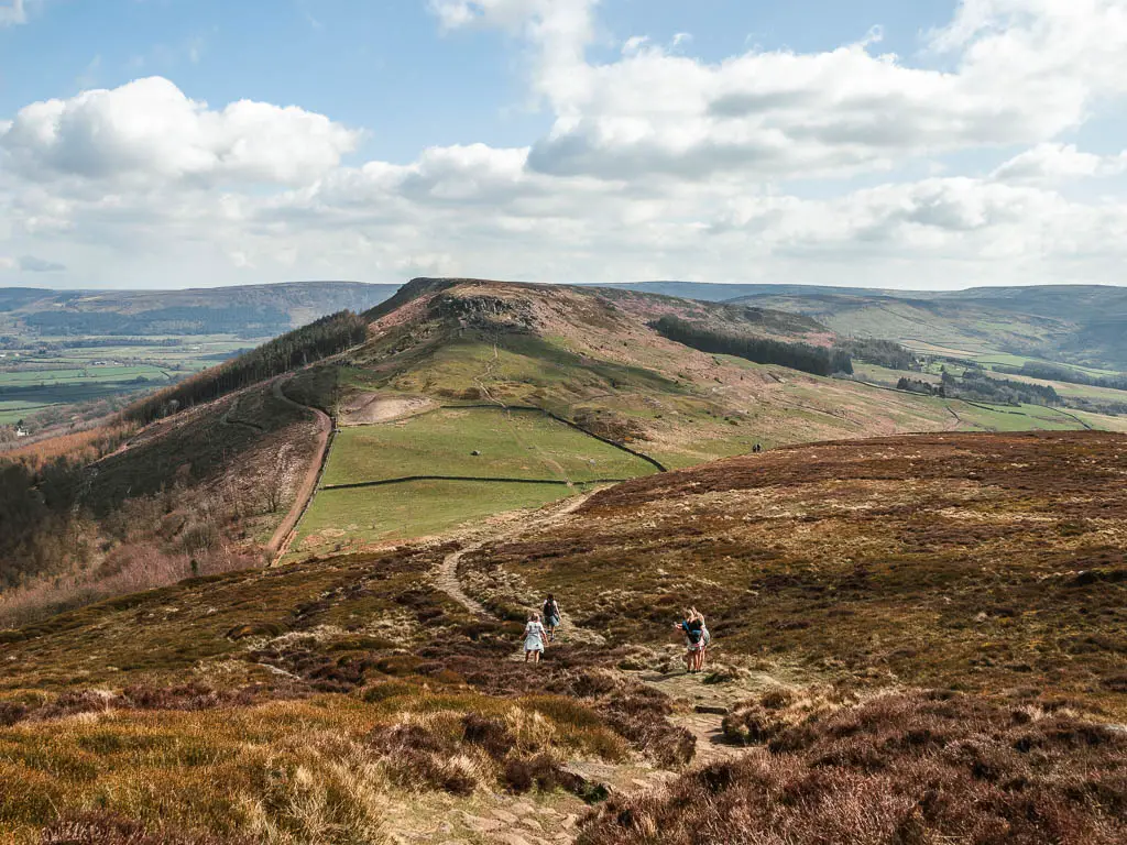 A trail with a few people walking along it, leading downhill into the valley, and a hill rising up on the other side, on the way to the Wainstones.