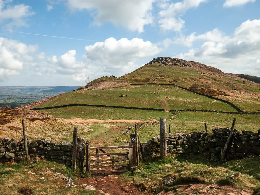 A wooden gate in the stone wall with a green grass hill rising up on the other side, on the walk towards the Wainstones.