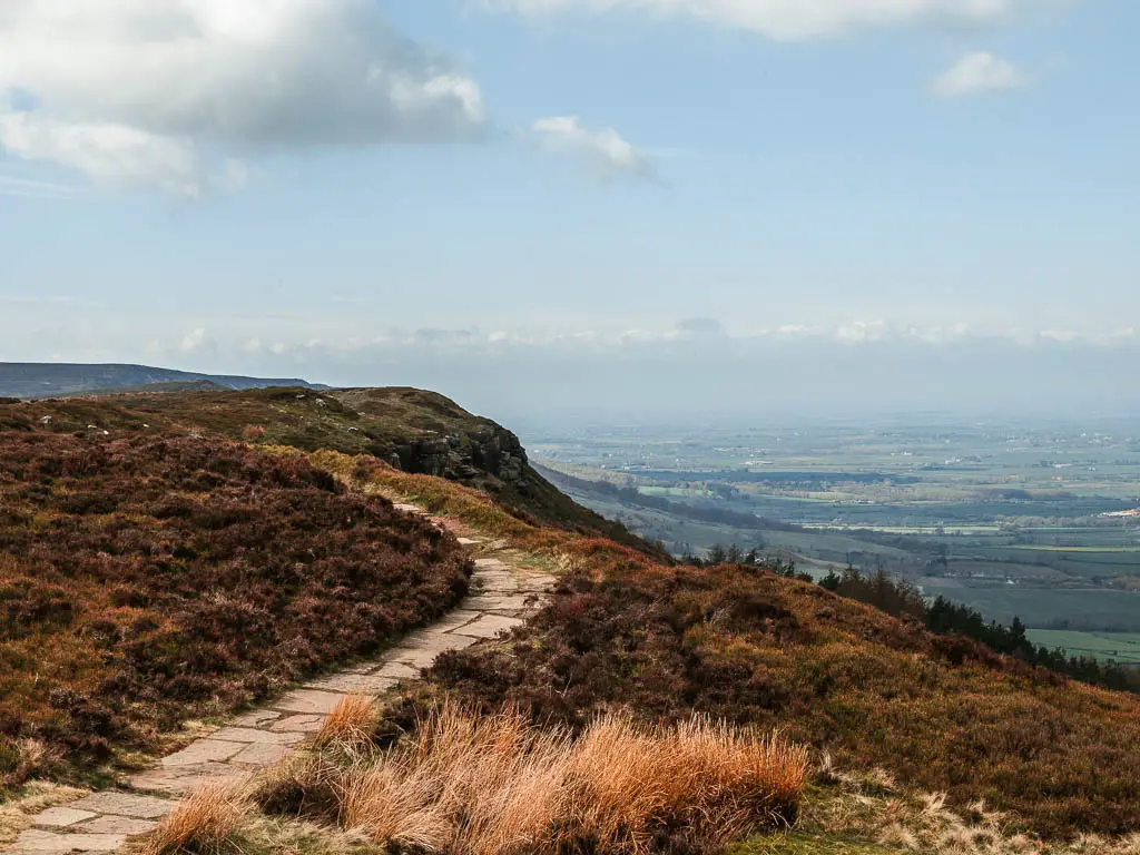 A stone paved trail curving along the end of the top of the hill, surrounded by brown heather, with a view down into the valley below on the right.