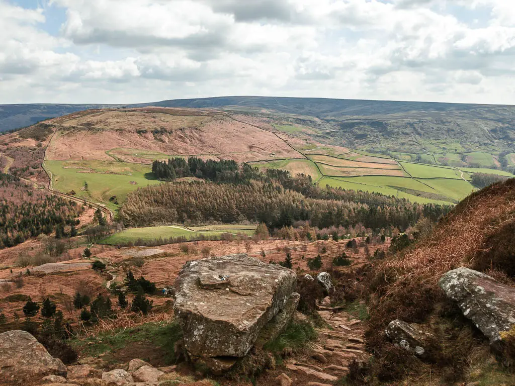 A stone paved trail leading downhill, through some rocks, with a vast view down into the valley filled with fields and masses of trees, and a hill rising up on the other side, on the walk away from the Wainstones.