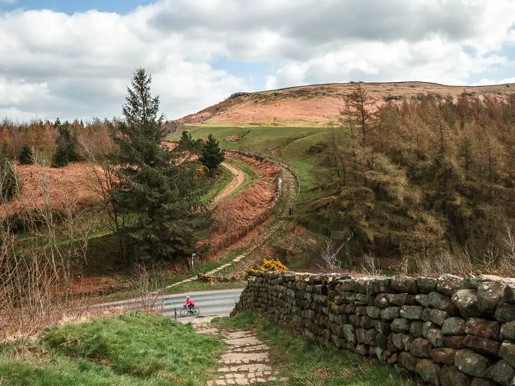 A stone paved trail leading down to the road, with a stone wall on the left. There is a hill rising up on the other side of the road with a trail running up it. There is a cyclist with a red jacket on the road.