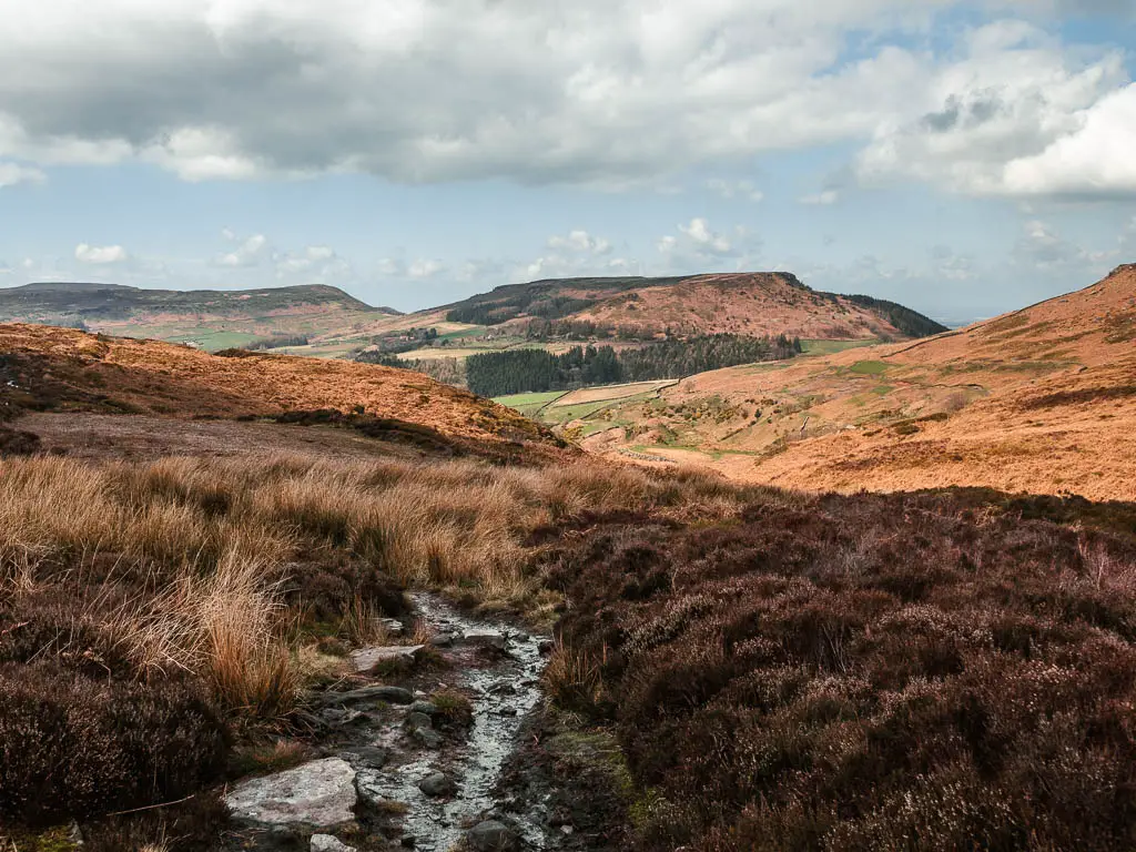 A wet strip of trail surround by brown heather and hay grass, with a view ahead to the valley and hills, with the sun shining down on them.