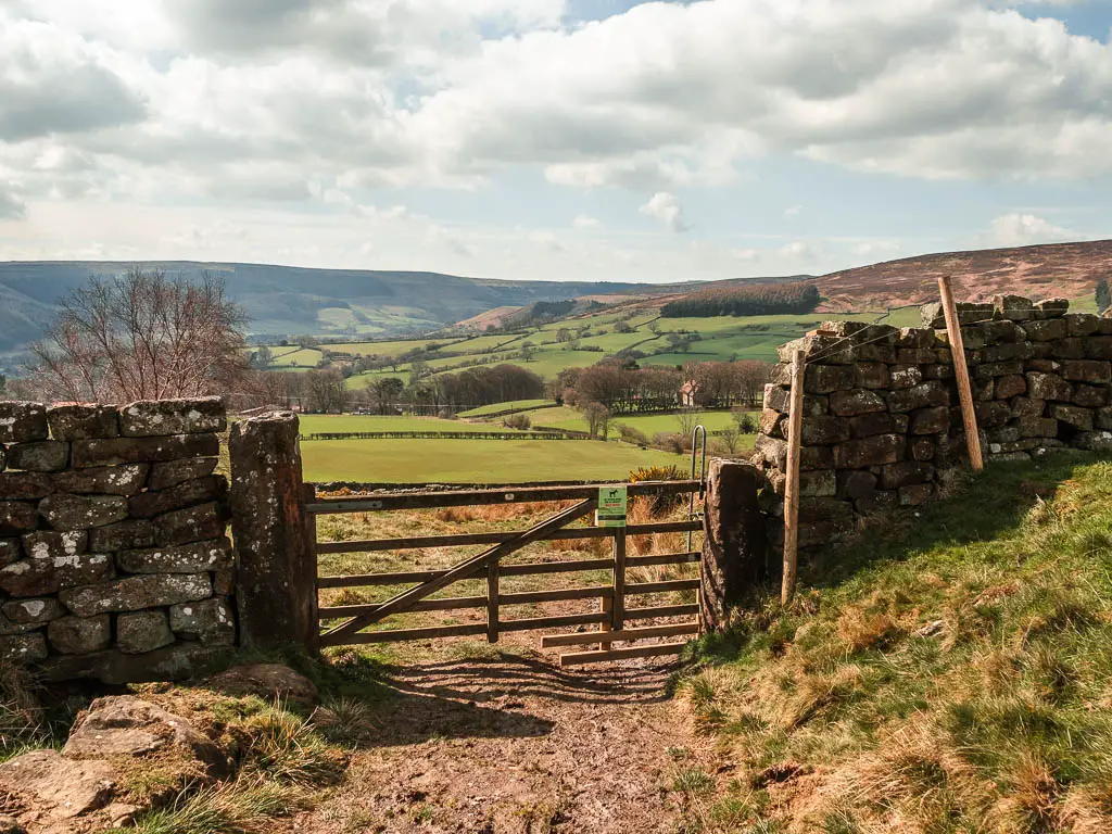 A wooden fence in the stone wall, with a view to green grass fields and hills on the other side in the distance. 