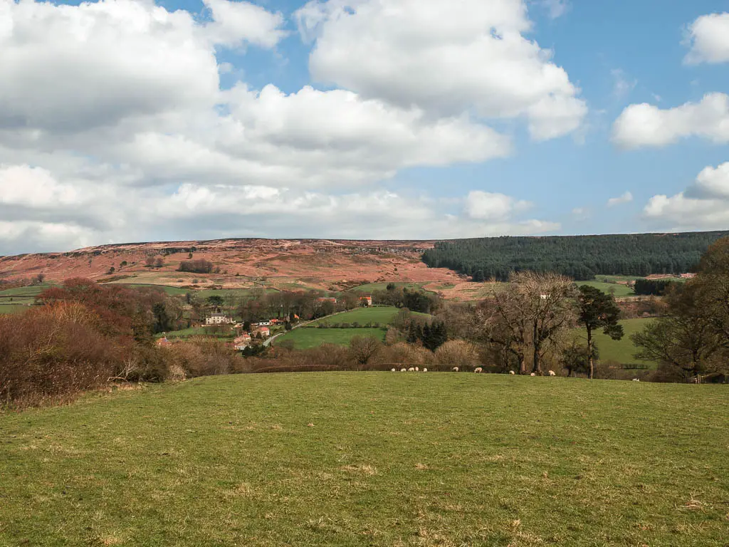 Looking down and across the hill of neatly cut green grass, with ground of trees below and a small group of houses at the bottom. There is a hill rising up on the other side.