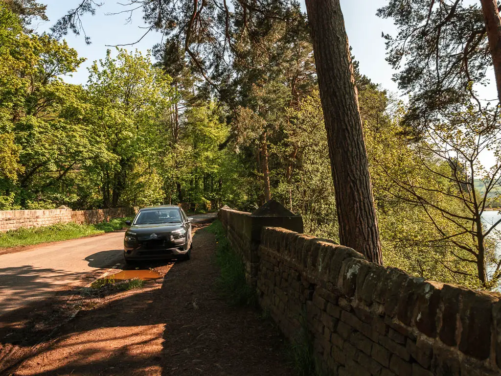 A stone wall on the right, and road on the left, with a black car parked for Agden Reservoir on the side.